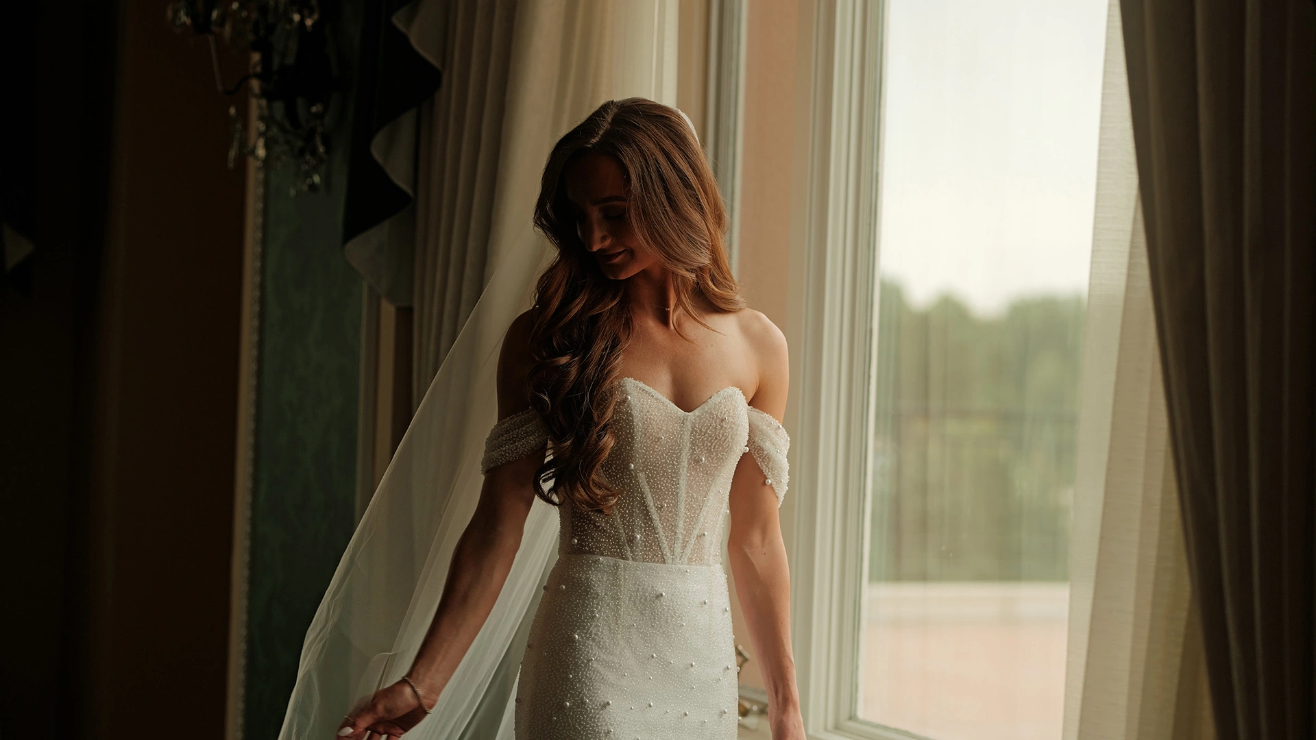 Bride in elegant beaded gown standing by window, editorial wedding portrait